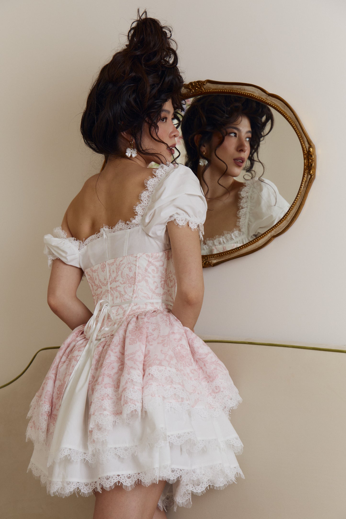 A woman with an elaborate dark hair bun poses in front of a vintage gold-framed mirror, wearing a white puff sleeve mini dress with lace accents.